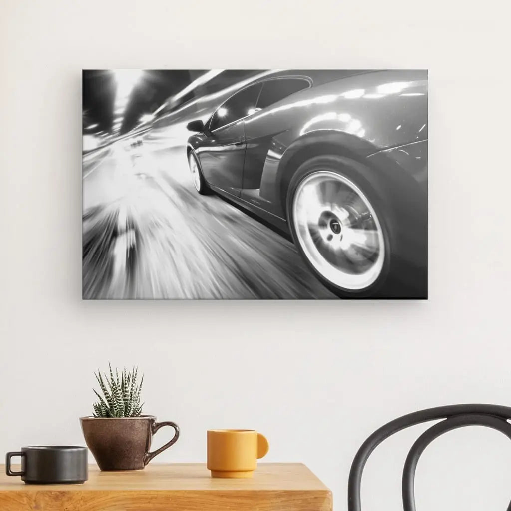 Photographie en noir et blanc du Tableau Voiture Autoroute Vitesse Noir et Blanc, accroché sur un mur blanc au-dessus d'une table en bois clair avec un petit cactus, une tasse marron et une tasse jaune. Une chaise noire est partiellement visible à droite.