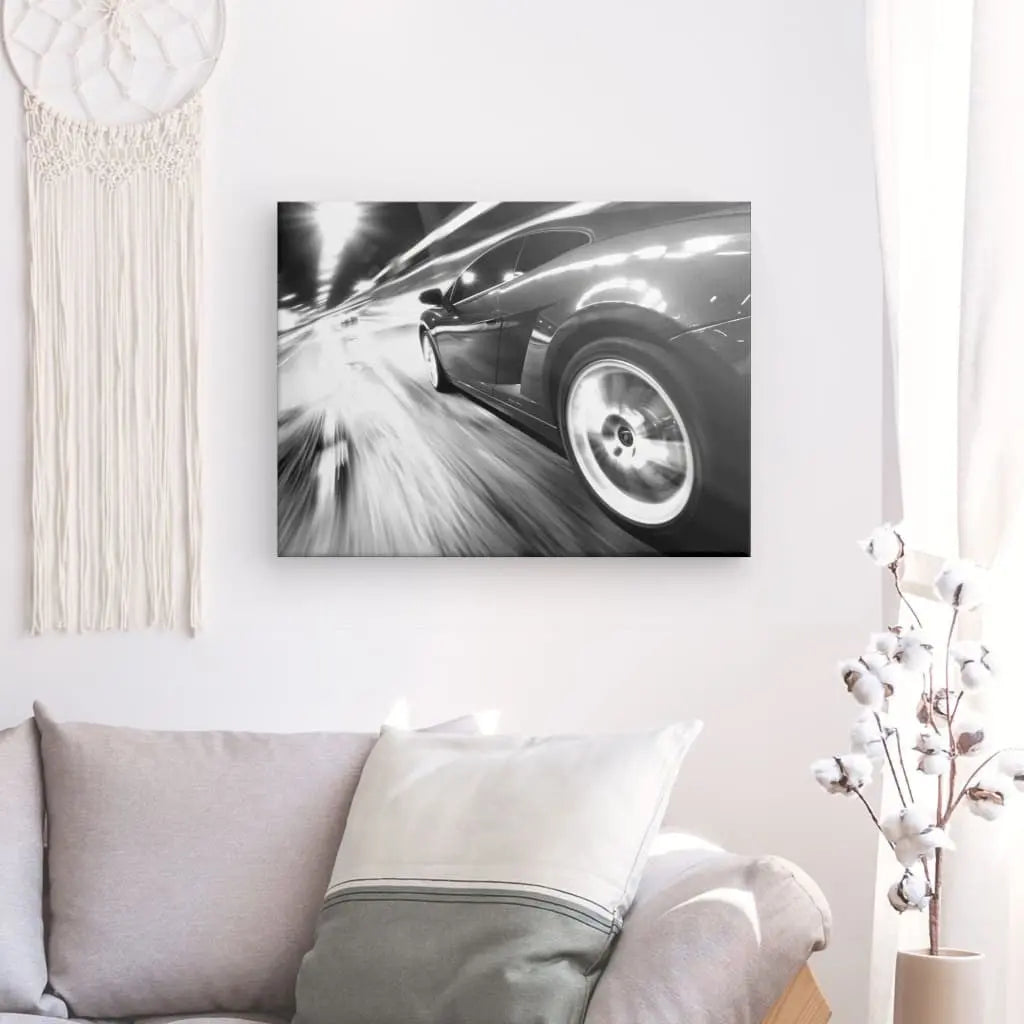 Salon comprenant un canapé gris avec un coussin blanc et gris, un Tableau Voiture Autoroute Vitesse Noir et Blanc mural, une pièce décorative suspendue en macramé et un vase blanc avec des branches en coton.