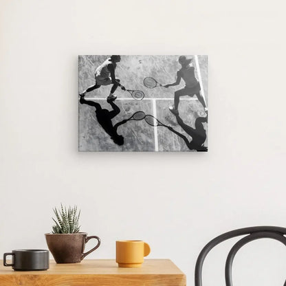 Un Tableau Cours Tennis Doublette Noir et Blanc représentant deux personnes jouant au tennis est accroché sur un mur blanc au-dessus d'une table en bois avec une plante en pot et deux tasses. Une chaise noire est placée à côté de la table, complétant la décoration intérieure élégante.