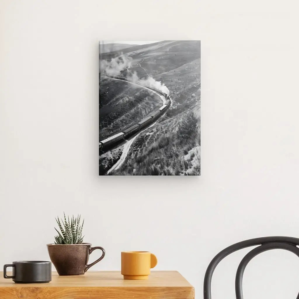 Le Tableau Collines Far West Train Vapeur Noir et Blanc, une photographie en noir et blanc d'un train, est présenté sur une toile tendue à la main accrochée à un mur blanc au-dessus d'une table en bois avec une petite plante en pot et deux mugs, l'un marron et l'autre jaune. La scène est complétée par la présence d'une chaise noire. La superbe définition d'impression utilisant des encres HP Latex renforce le charme intemporel de cette imagerie classique.