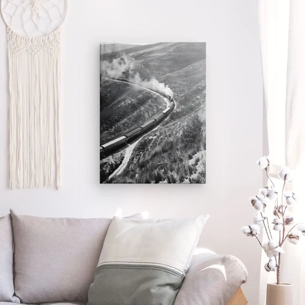 Le Tableau Collines Far West Train Vapeur Noir et Blanc, une photographie en noir et blanc d'un train à vapeur sur une voie sinueuse, est accroché sur un mur blanc au-dessus d'un canapé beige avec un coussin vert. La superbe définition de l'impression est évidente, rendue possible grâce aux encres HP Latex, et la toile tendue à la main ajoute une touche élégante au décor. Une pièce décorative blanche se trouve à proximité.