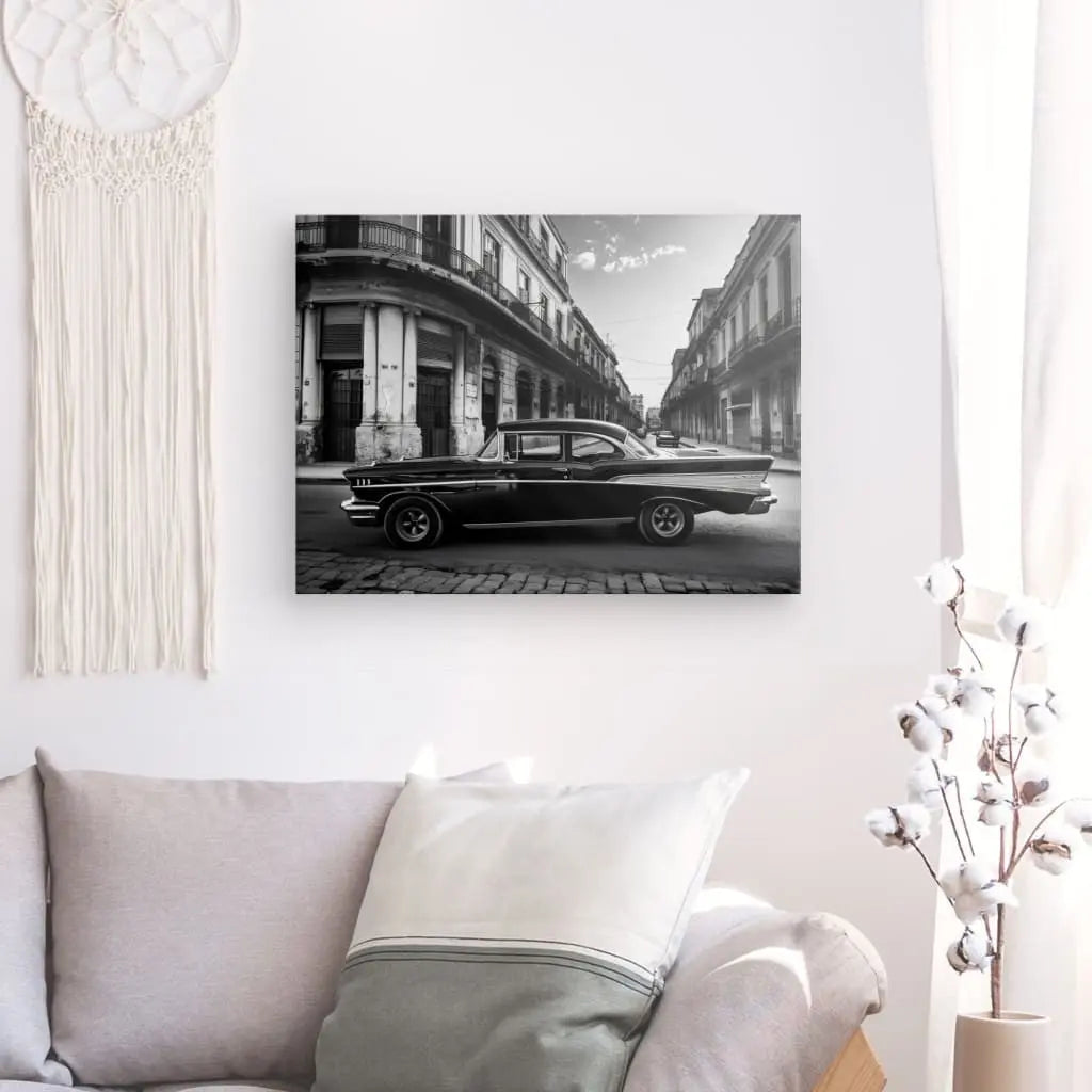 Le Tableau Voiture Cubaine Rue Noir et Blanc, reproduction en noir et blanc d'une voiture ancienne dans une rue de la ville, encadrée en bois d'épicéa, est accroché sur un mur blanc au-dessus d'un canapé clair avec des coussins. Une tenture murale en macramé se trouve à gauche et une plante en pot à droite.