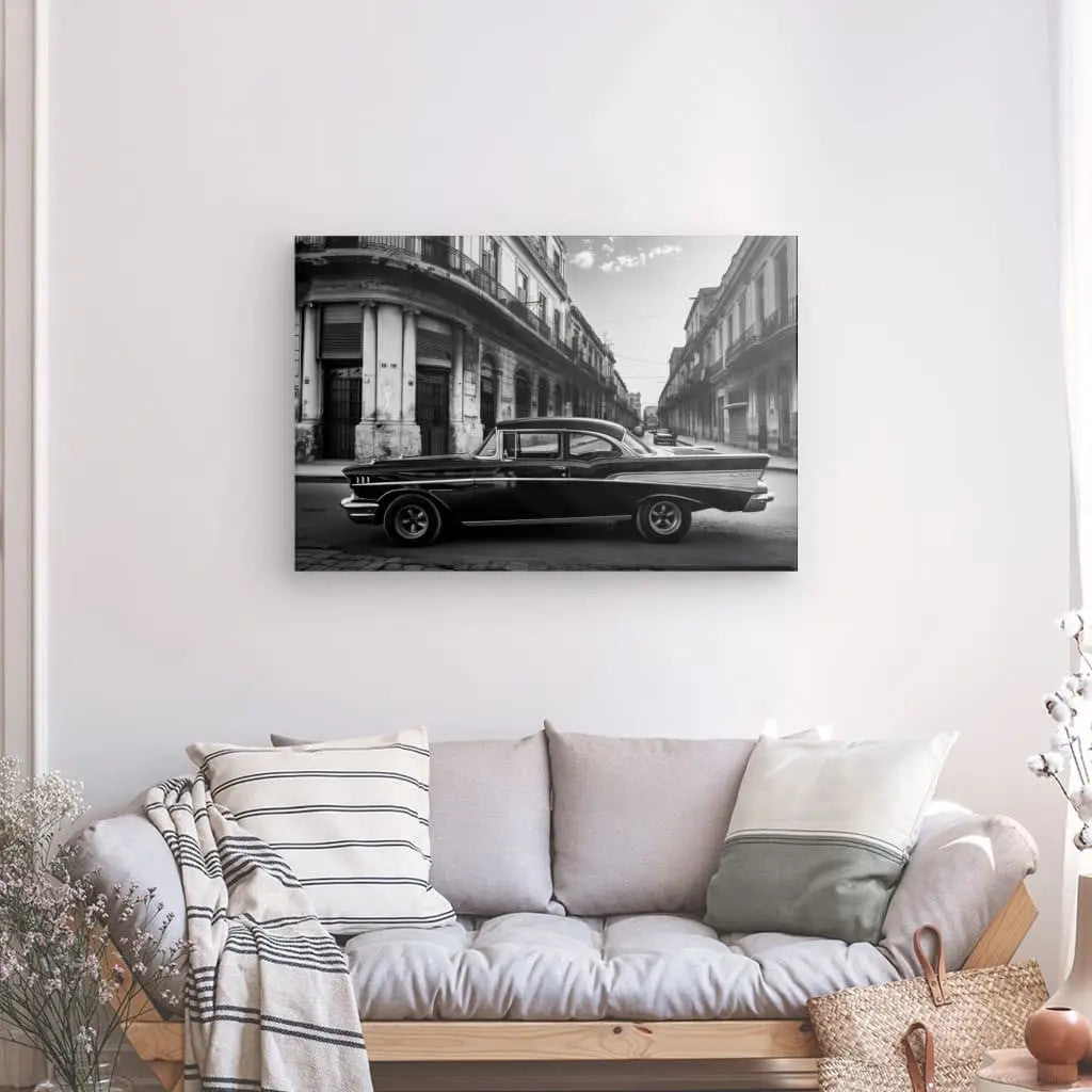 Le Tableau Voiture Cubaine Rue Noir et Blanc, composé de reproductions photographiques en noir et blanc d'une voiture ancienne dans une rue de la ville, est suspendu au-dessus d'un canapé beige orné de coussins rayés et unis. Un panier contenant un plaid et un feuillage vert est placé à gauche du canapé, mettant en valeur ce classique du design polyvalent.