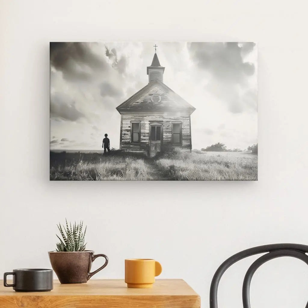 Une photographie en noir et blanc intitulée « Tableau Eglise Campagne Nuage et Soleil Monochrome » représente une personne debout près d'une vieille église patinée par les intempéries dans un champ. L'image est tendue à la main sur une toile et montée sur un mur blanc au-dessus d'une table en bois ornée d'une chaise, de tasses et d'une plante en pot.