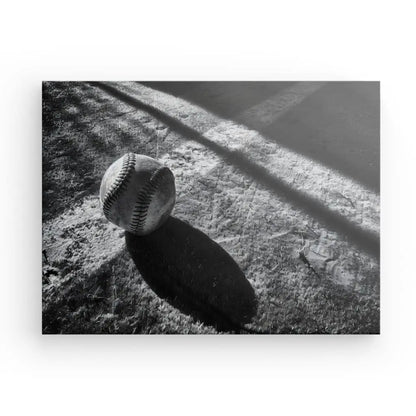 Le Tableau Balle Baseball Terrain Noir et Blanc capture une balle de baseball posée sur le sol, projetant une ombre et se trouvant à côté d'une ligne marquée sur une surface inégale, évoquant la sensation de reproductions de photos vintage en noir et blanc.