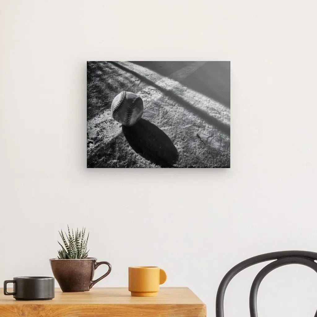 Reproduction photographique en noir et blanc d'une balle de baseball projetant une ombre sur une surface, montée sur un mur au-dessus d'une table avec une plante en pot et deux tasses, et imprimée à l'aide d'encres latex HP, intitulée « Tableau Balle Baseball Terrain Noir et Blanc ».