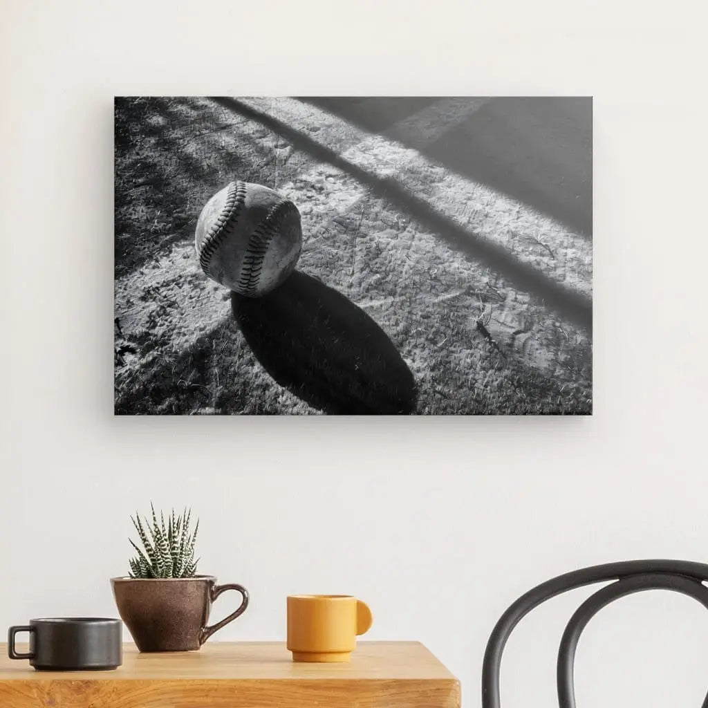 Photographie en noir et blanc d'une balle de baseball sur une surface texturée, reproduite avec des encres HP Latex et affichée sur un mur au-dessus d'une petite table avec une plante, une tasse et un mug. La photographie est intitulée "Tableau Balle Baseball Terrain Noir et Blanc".