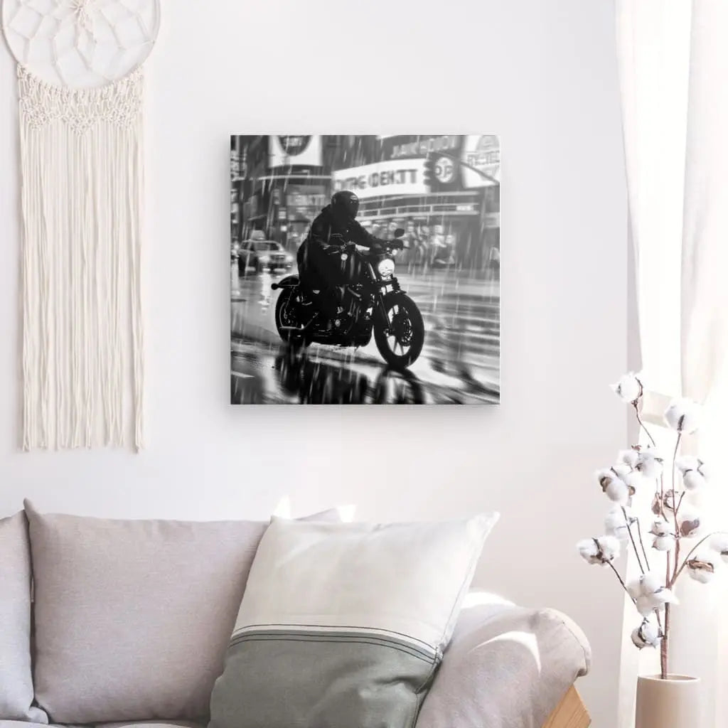 Le Tableau Motard Ville Pluvieuse Nuit Noir et Blanc, une photo encadrée en noir et blanc d'un motocycliste roulant sous la pluie, imprimée avec des encres HP Latex sur une toile blanche brillante à texture fine, est montée sur un mur blanc au-dessus d'un canapé beige avec une tenture murale à franges et une plante décorative.