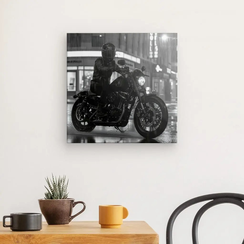 Le Tableau Moto Virée Urbaine Nocturne Monochrome, une reproduction photographique en noir et blanc d'un motocycliste la nuit, est monté sur un mur blanc au-dessus d'une table avec un cactus, une tasse marron, une tasse jaune et une chaise noire. L'image ressort grâce aux encres HP Latex vibrantes.