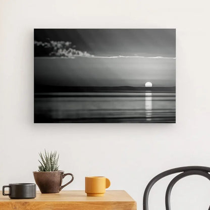 Le Tableau Coucher de Soleil Mer Noir et Blanc, une reproduction photographique en noir et blanc d'un coucher de soleil sur un plan d'eau, est affiché sur un mur blanc au-dessus d'une table en bois ornée d'une plante en pot et de deux tasses. Une chaise noire est partiellement visible à droite. Cette image, rehaussée d'encres HP Latex, capture parfaitement la beauté sereine du moment.