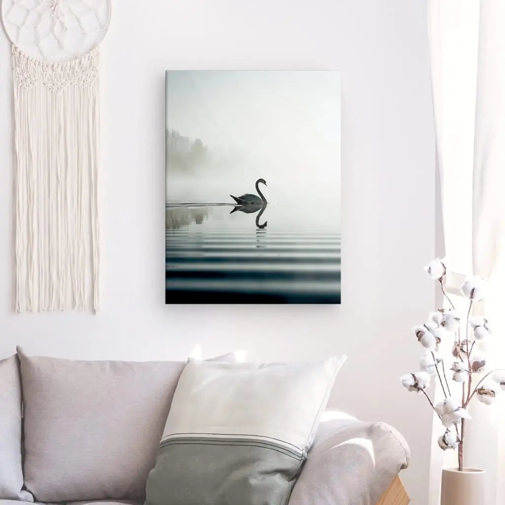 Un Tableau Cygne Lac Style Epuré, représentant un cygne flottant sur un lac brumeux, est accroché sur un mur blanc au-dessus d'un canapé gris clair dans une pièce de style épuré avec un décor suspendu blanc et une plante de coton blanche.