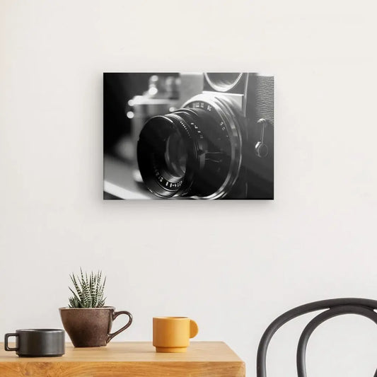 Un Tableau Photo Objectif Obturateur Noir et Blanc est accroché sur un mur blanc au-dessus d'une table en bois avec une plante en pot, une tasse marron et une tasse orange. Le bord d'une chaise est visible, ajoutant au charme de la décoration intérieure.