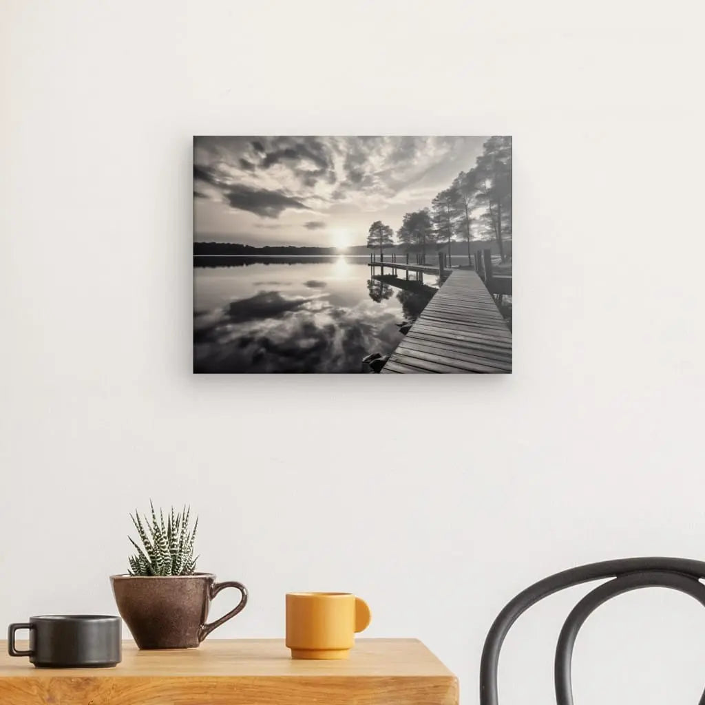 Un Tableau Ponton Lac Nature Noir et Blanc est accroché sur un mur blanc au-dessus d'une table en bois avec une plante en pot et deux tasses, à côté d'une chaise noire.