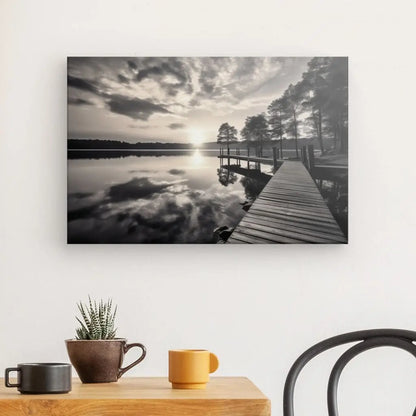 Un Tableau Ponton Lac Nature Noir et Blanc sur un mur représente un ponton dans un lac, s'étendant dans des eaux calmes sous un ciel nuageux, flanqué d'arbres. Ci-dessous, une table avec une petite plante et deux tasses est partiellement visible.