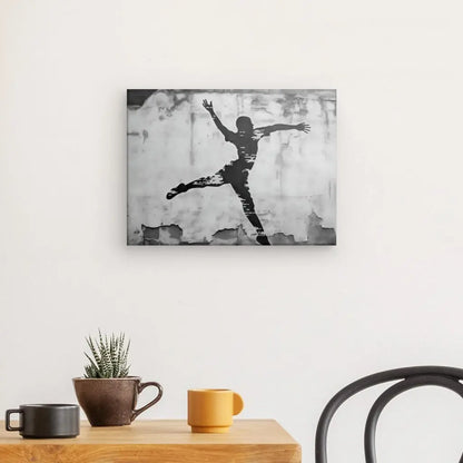 Oeuvre encadrée en noir et blanc représentant une silhouette de personne sautant sur un mur blanc au-dessus d'une table en bois avec une tasse marron et jaune, une plante en pot et une chaise noire. Le Tableau Basketteur Underground Tag Noir et Blanc ajoute une touche urbaine à la pièce, rappelant l'art de rue.