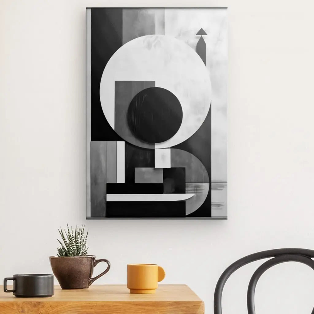 Art géométrique en noir et blanc sur un mur blanc au-dessus d'une table en bois avec plante en pot, tasse marron et tasse jaune. Une chaise noire est visible sur le côté droit, créant un Tableau Géométrique Abstrait Contrastes Cubisme Noir et Blanc moderne.