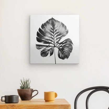 Une peinture monochrome représentant un Tableau Feuille Végétale Naturelle Noir et Blanc est accrochée à un mur au-dessus d'une table en bois avec une tasse marron, une tasse jaune et une petite plante en pot. Une chaise noire est partiellement visible, ajoutant à l'élégance naturelle de la scène.