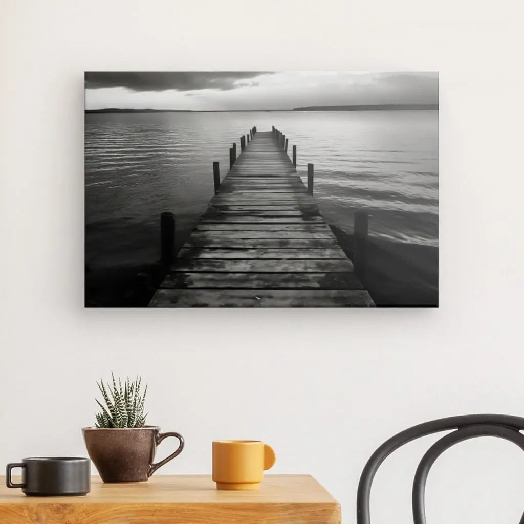 Tableau Ponton Lac en Bois Paysage Noir et Blanc imprimé d'une jetée en bois menant à un lac calme sous un ciel nuageux, affiché au-dessus du Tableau Ponton Lac en Bois Paysage Noir et Blanc avec une plante et une tasse.